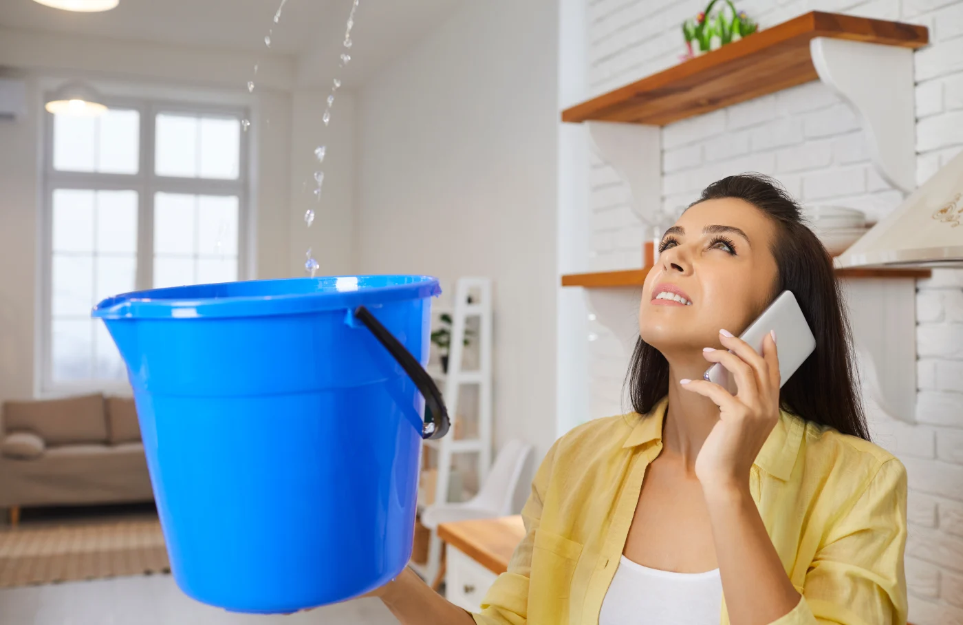 A homeowner holds a bucket under a roof leak while calling for help.