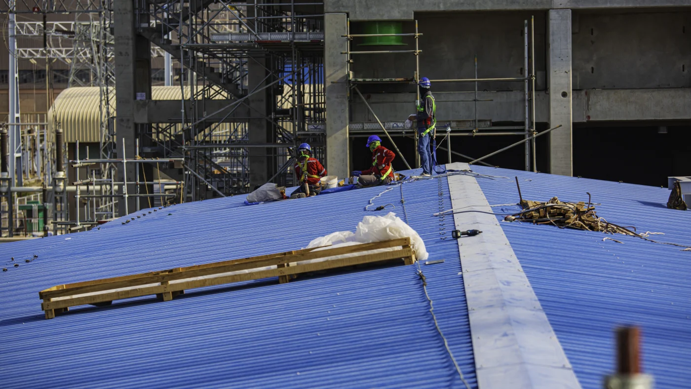 Construction crew in safety gear working on a large blue metal commercial roof, with scaffolding and industrial structures in the background.