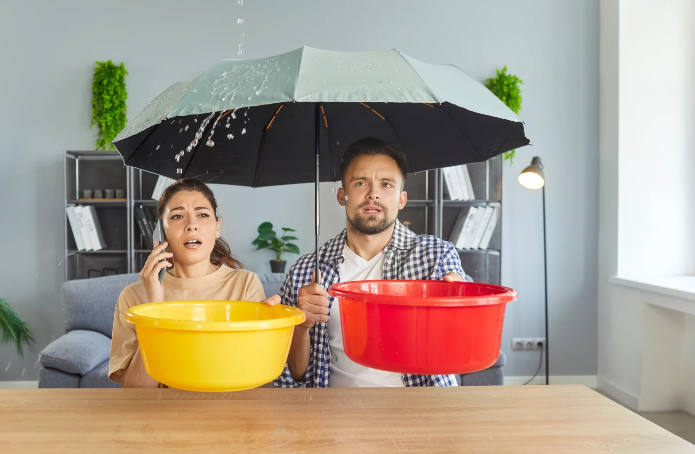 A couple sits under an umbrella while holding buckets to catch a roof leak