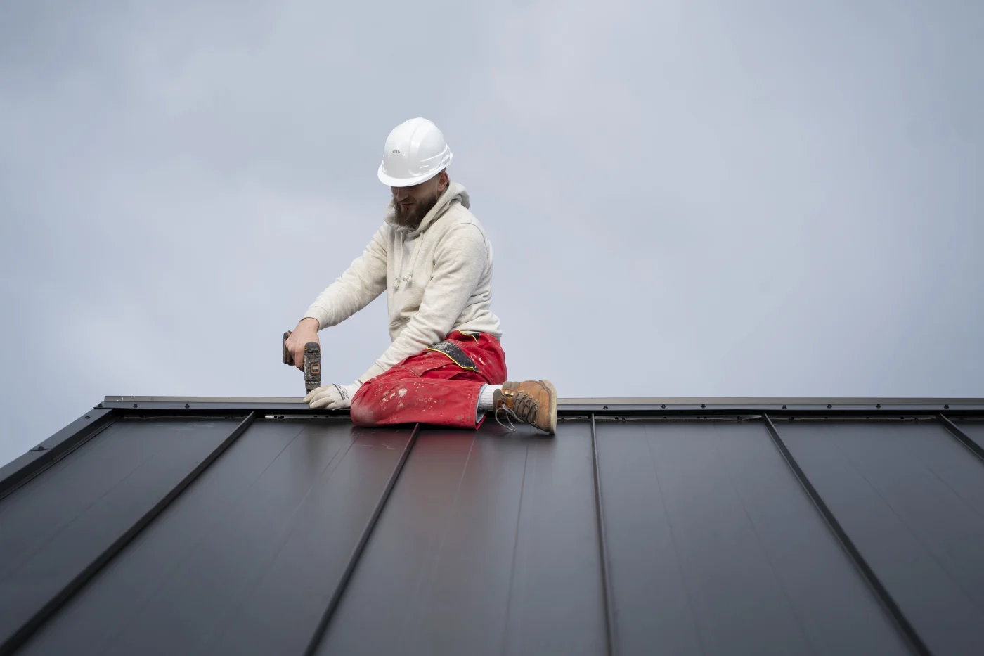 Roofer in safety gear and hard hat sitting on a black metal roof, using a power drill to secure panels under a cloudy sky.