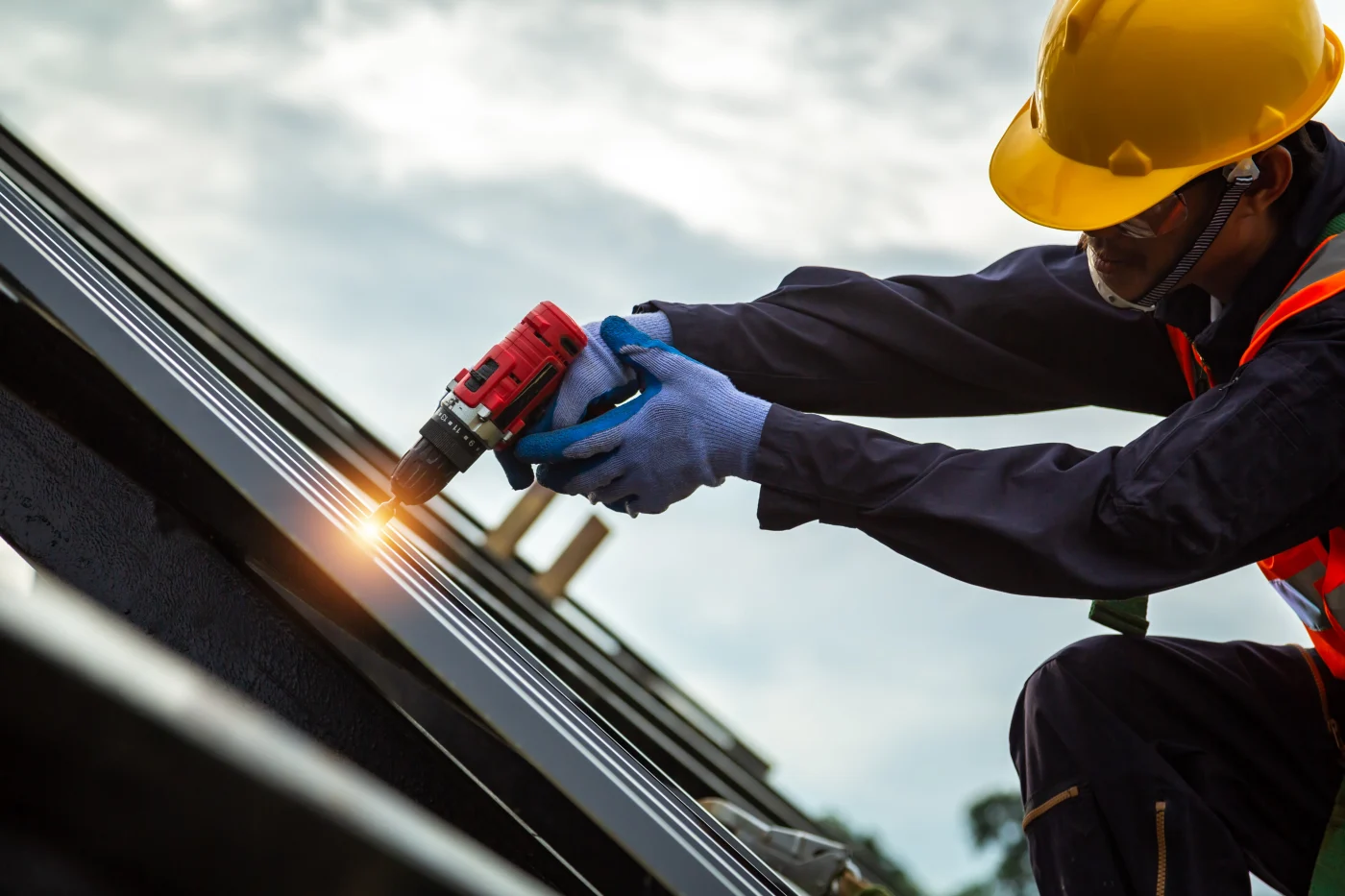 Construction worker in safety gear using a red power drill to fasten a metal roof panel, wearing gloves, safety glasses, and a yellow hard hat.