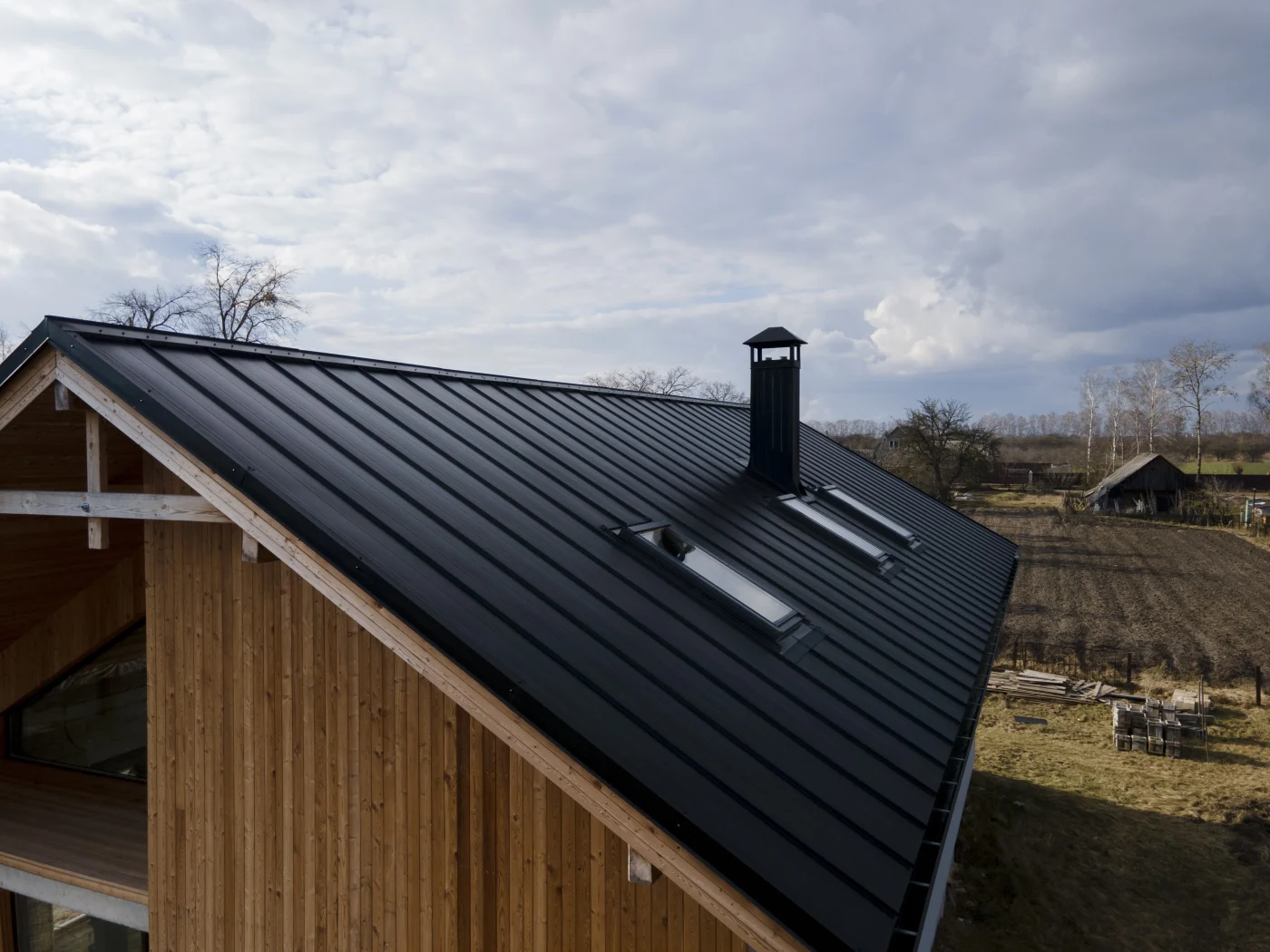 A modern house with a sleek black standing seam metal roof, skylights, and chimney, set against a rural landscape with cloudy sky and farmland