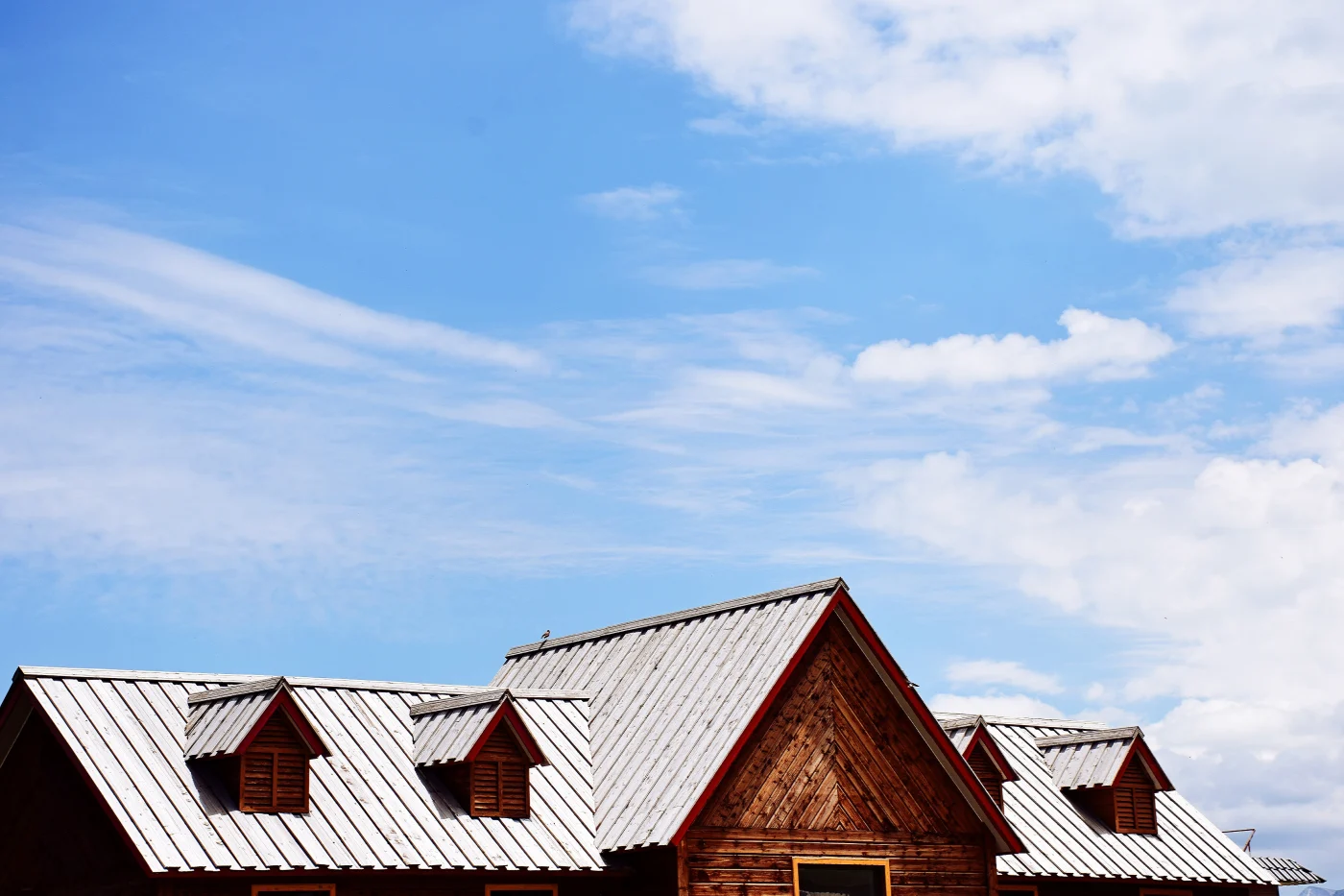 Wooden building with a metal gable roof and dormer windows under a bright blue sky with scattered clouds.