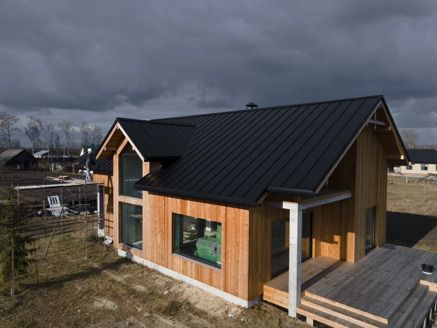 A modern wooden house with large windows and a sleek black metal roof is under construction.