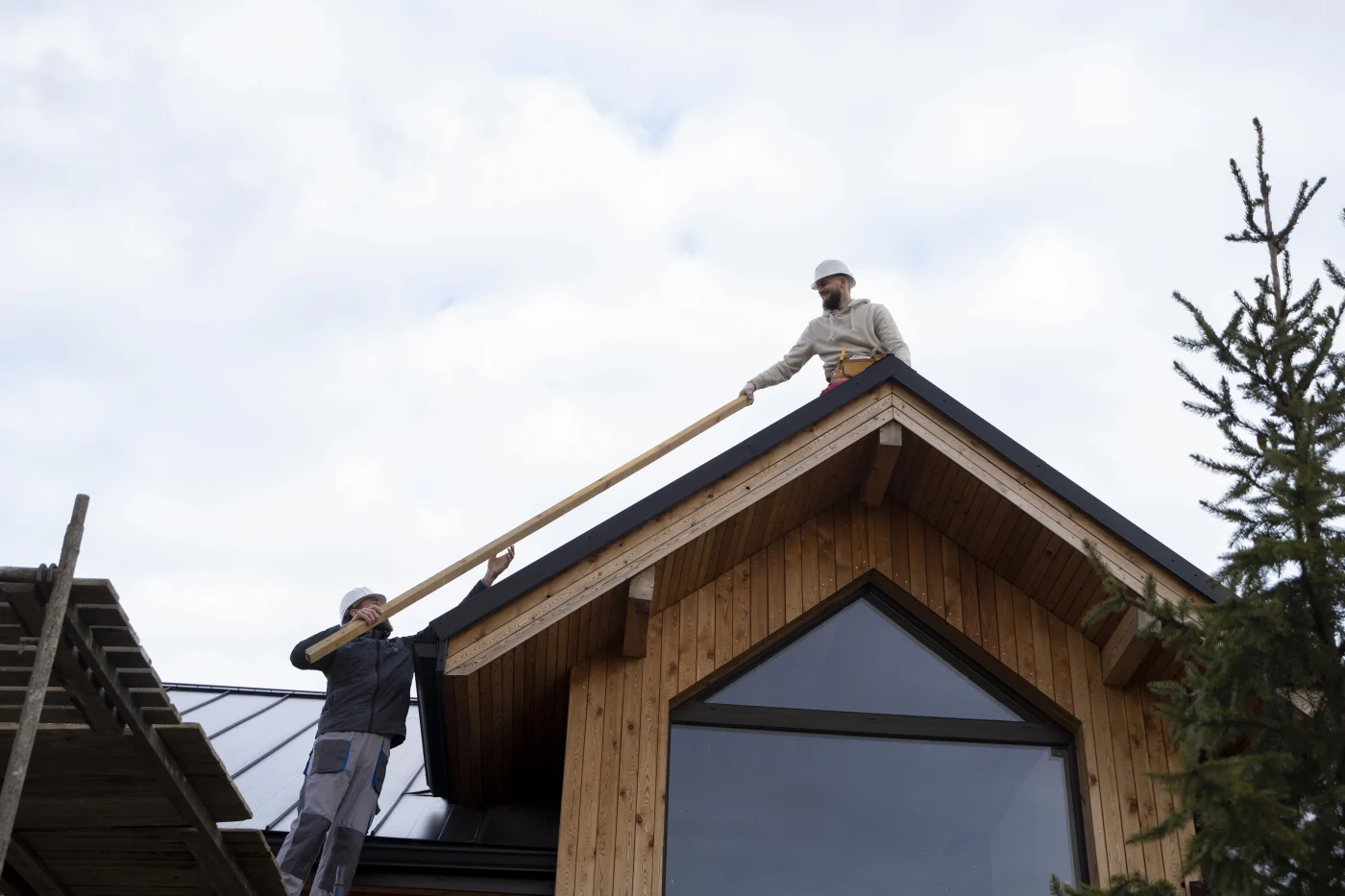 Two roofers in safety gear installing wooden beams on a steep residential roof, one standing on scaffolding and the other positioned at the roof peak.