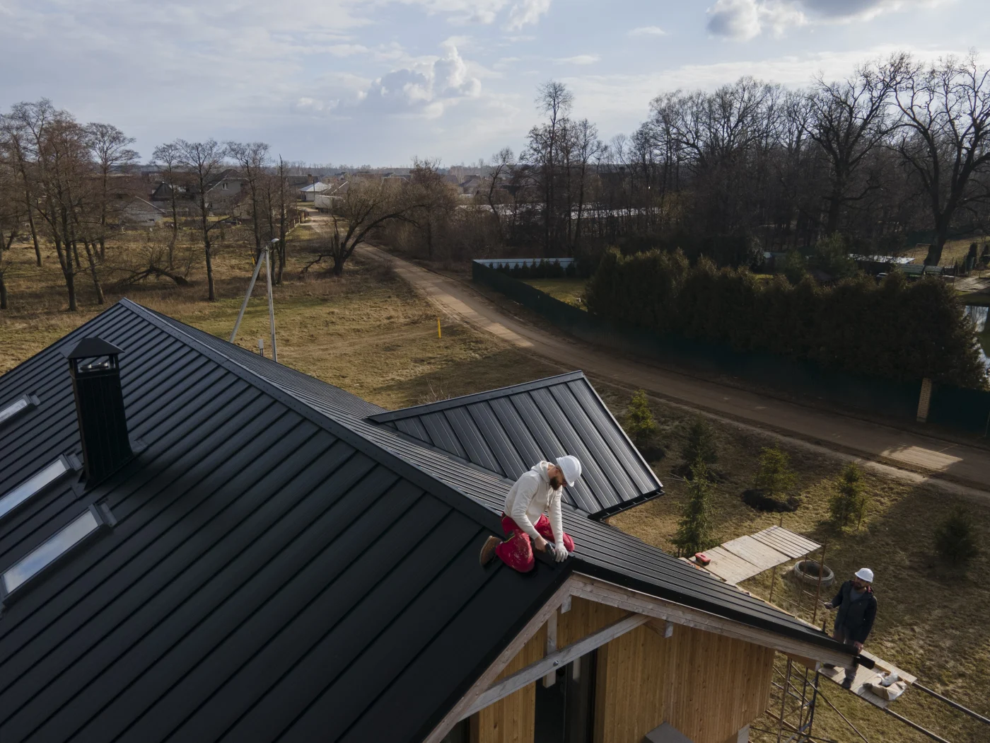 Roofing contractors installing black metal panels on a modern home roof, with safety gear and scaffolding under a cloudy sky in a rural neighborhood.