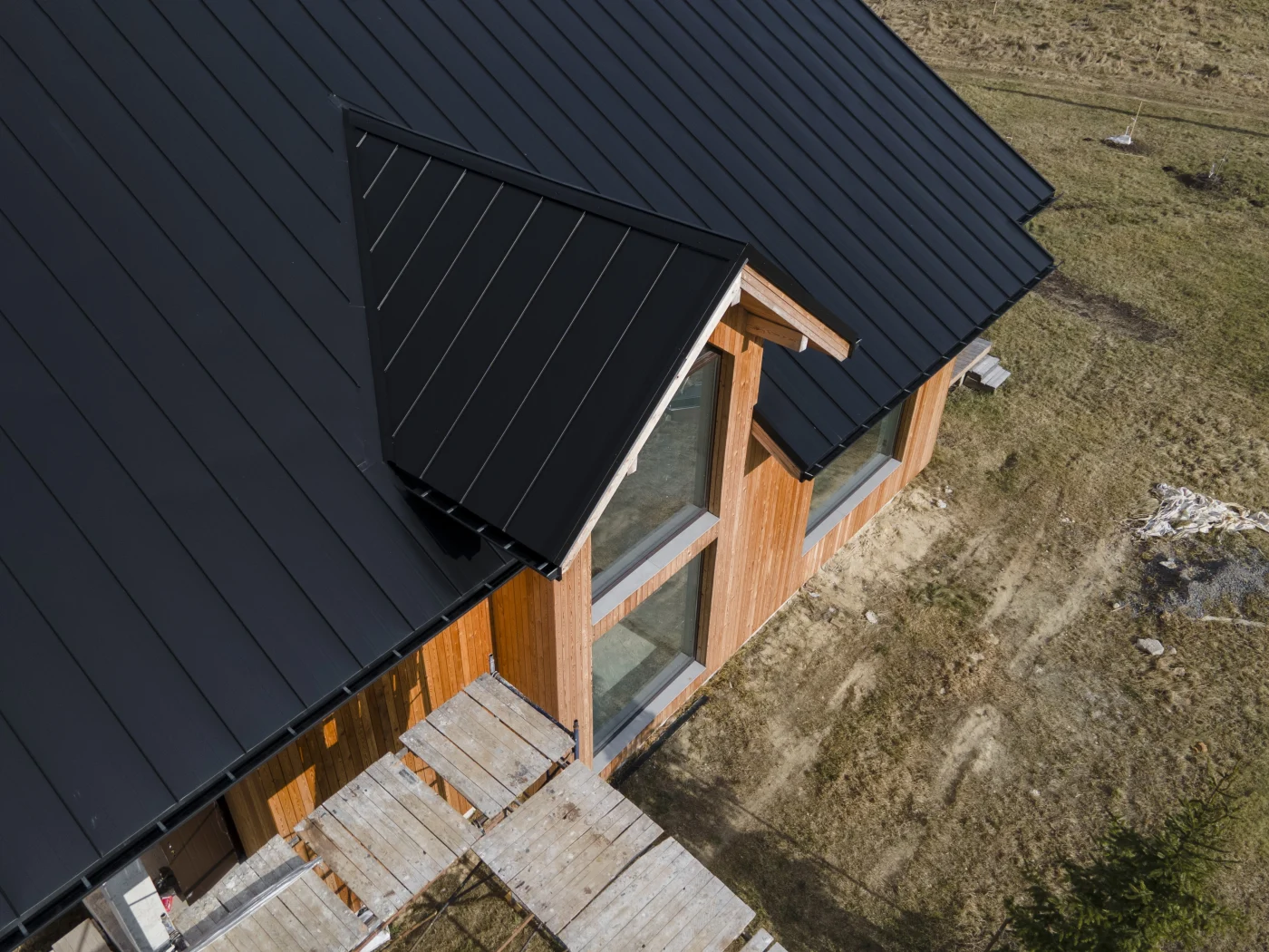 Modern wooden house with sleek black standing seam metal roofing, featuring skylights and a central chimney, set against a cloudy rural landscape.