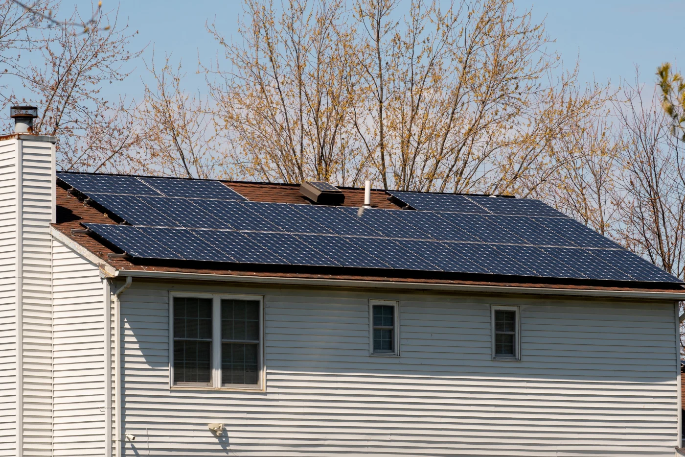Residential home with white siding and a roof covered in dark solar panels, surrounded by leafless trees under a bright sky.