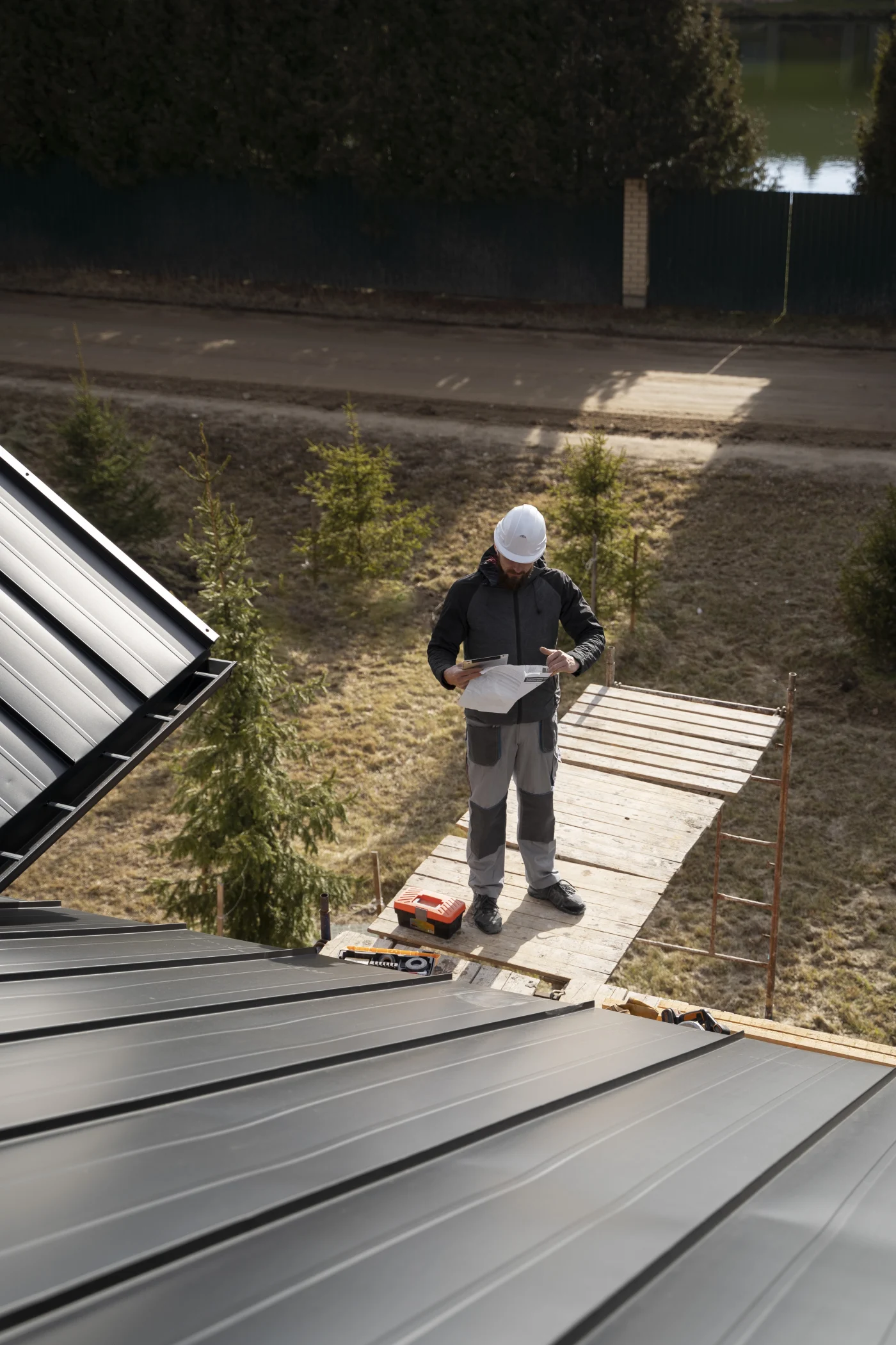 Construction worker wearing a hard hat standing on a wooden scaffold beside a metal roof installation, reviewing building plans with tools and equipment nearby.