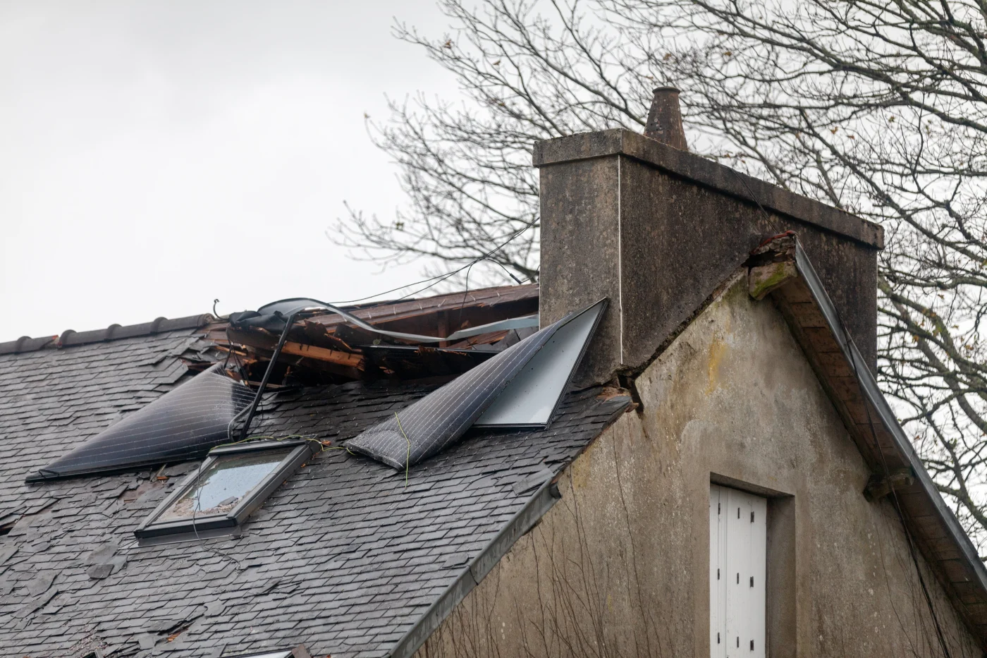Effective Storm Damage Roof Restoration | Longmont, CO | Columbine Roofing and Solar A storm-damaged roof with broken shingles, shattered skylight glass, and partially detached solar panels beside a worn chimney on an older home.