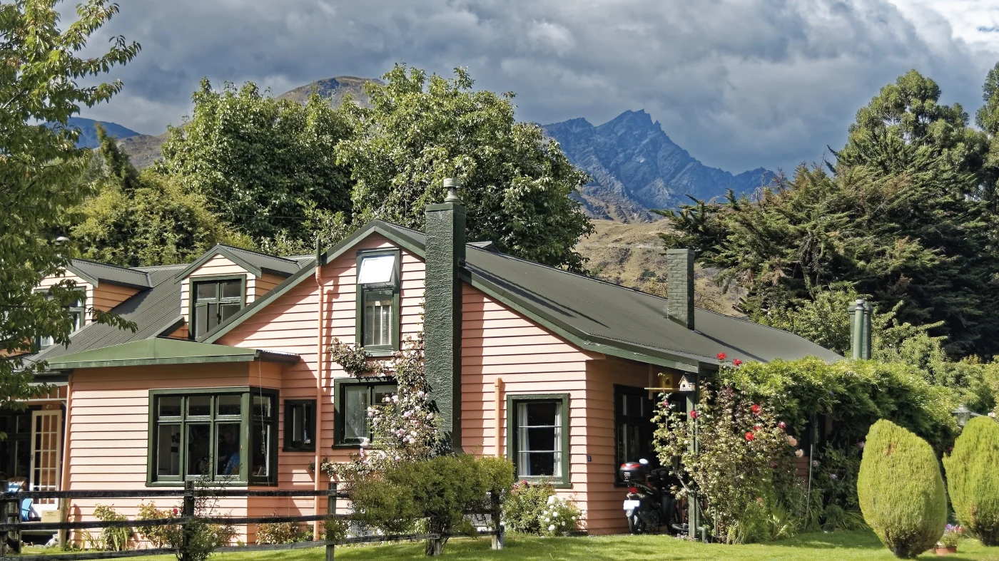 Charming peach-colored home with green trim and a dark metal roof, surrounded by lush trees and gardens with mountains rising in the background under cloudy skies.