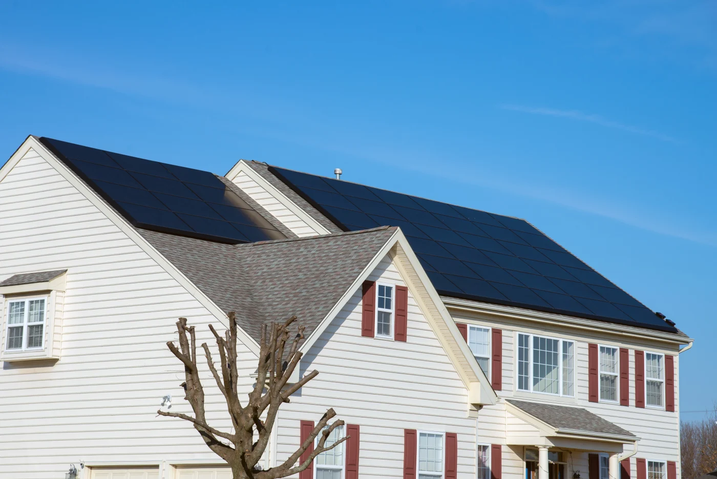 Two-story suburban home with large black solar panels installed across multiple roof sections under a clear blue sky.