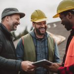 Two roofing professionals wearing hard hats review documents with a customer, smiling and collaborating to ensure quality workmanship and clear communication