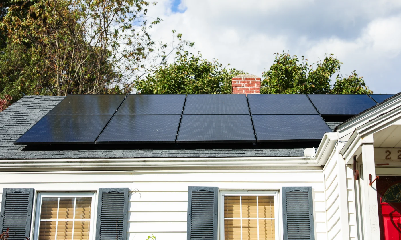 Single-story white house with dark shutters and a gray shingle roof topped with a neat row of black solar panels in front of leafy trees.
