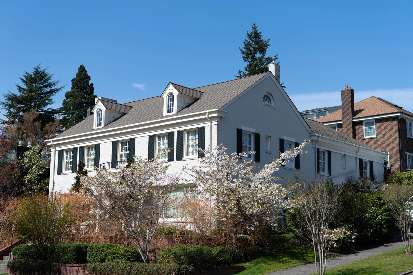 Elegant two-story white brick home with a tan asphalt shingle roof and black shutters, surrounded by blooming trees and lush landscaping on a sunny day