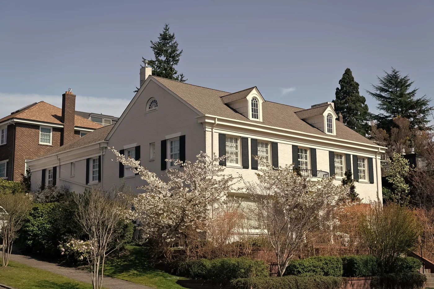 Two-story cream-colored brick home with a tan asphalt shingle roof, black shutters, and blossoming trees in the front yard.