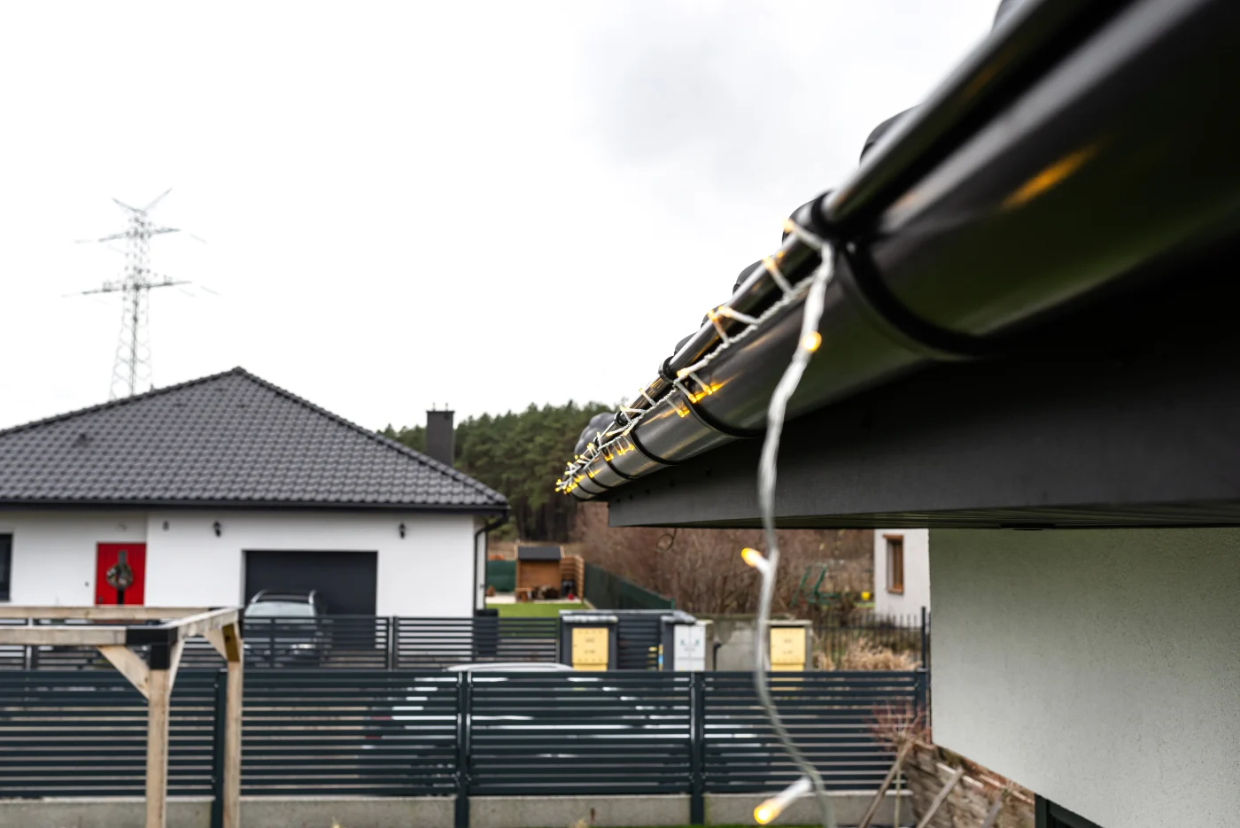 Close-up of a dark metal gutter on a modern home with string lights draped along the edge, overlooking a fenced yard and a neighboring white house.