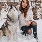 A woman in a white winter jacket kneels in the snow beside a husky dog, smiling outdoors in a snowy residential setting.