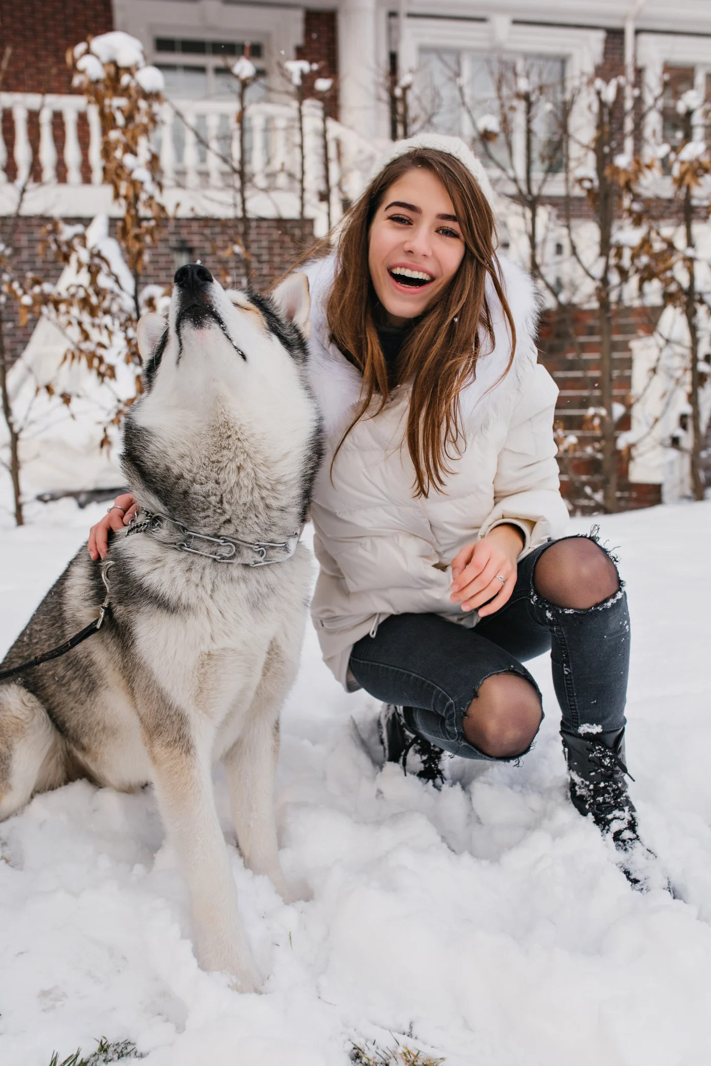 A woman in a white winter jacket kneels in the snow beside a husky dog, smiling outdoors in a snowy residential setting.