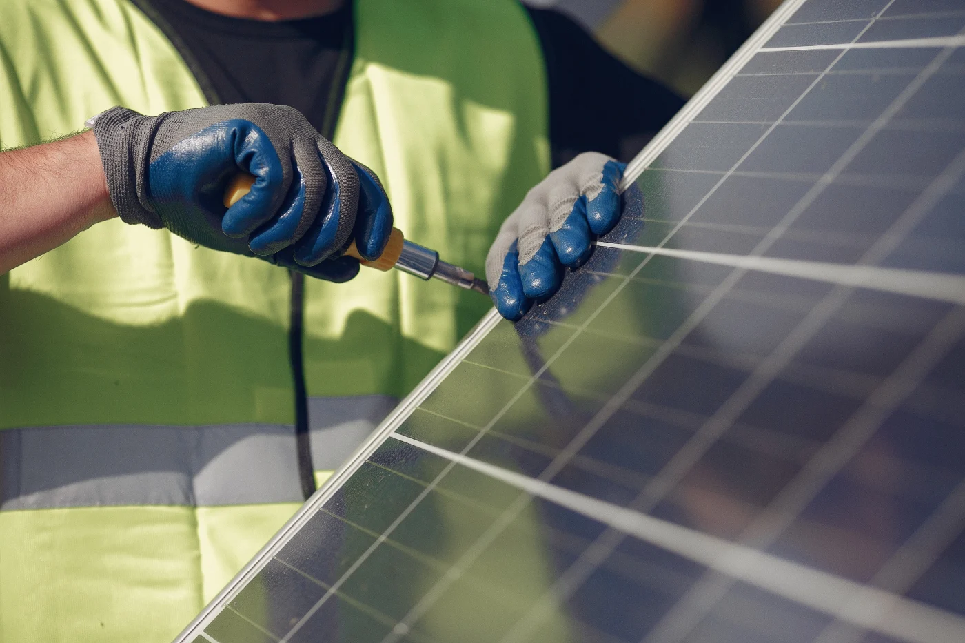 Close-up of a solar installer in a neon safety vest tightening hardware on a solar panel with a screwdriver, wearing blue and gray protective gloves.