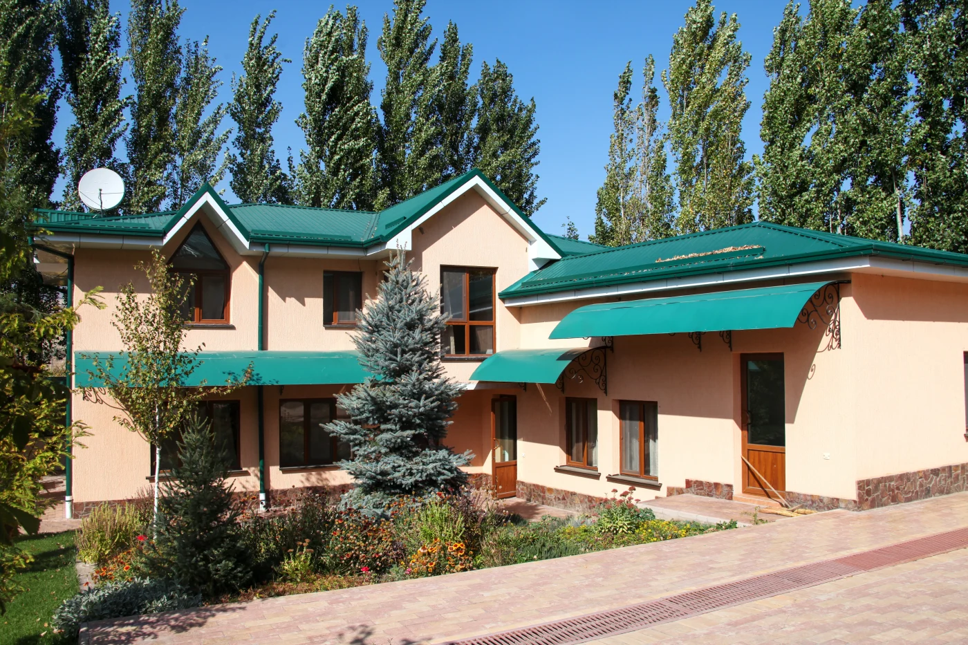 Perfect Residential Roof Installation | Boulder, CO | Columbine Roofing & Solar Two-story stucco home with a green metal roof, matching green awnings, and wood-framed windows, surrounded by trees and colorful garden beds.
