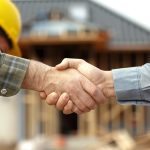 Two people shake hands at a residential construction site, symbolizing trust and professionalism, with a home under construction visible in the background.
