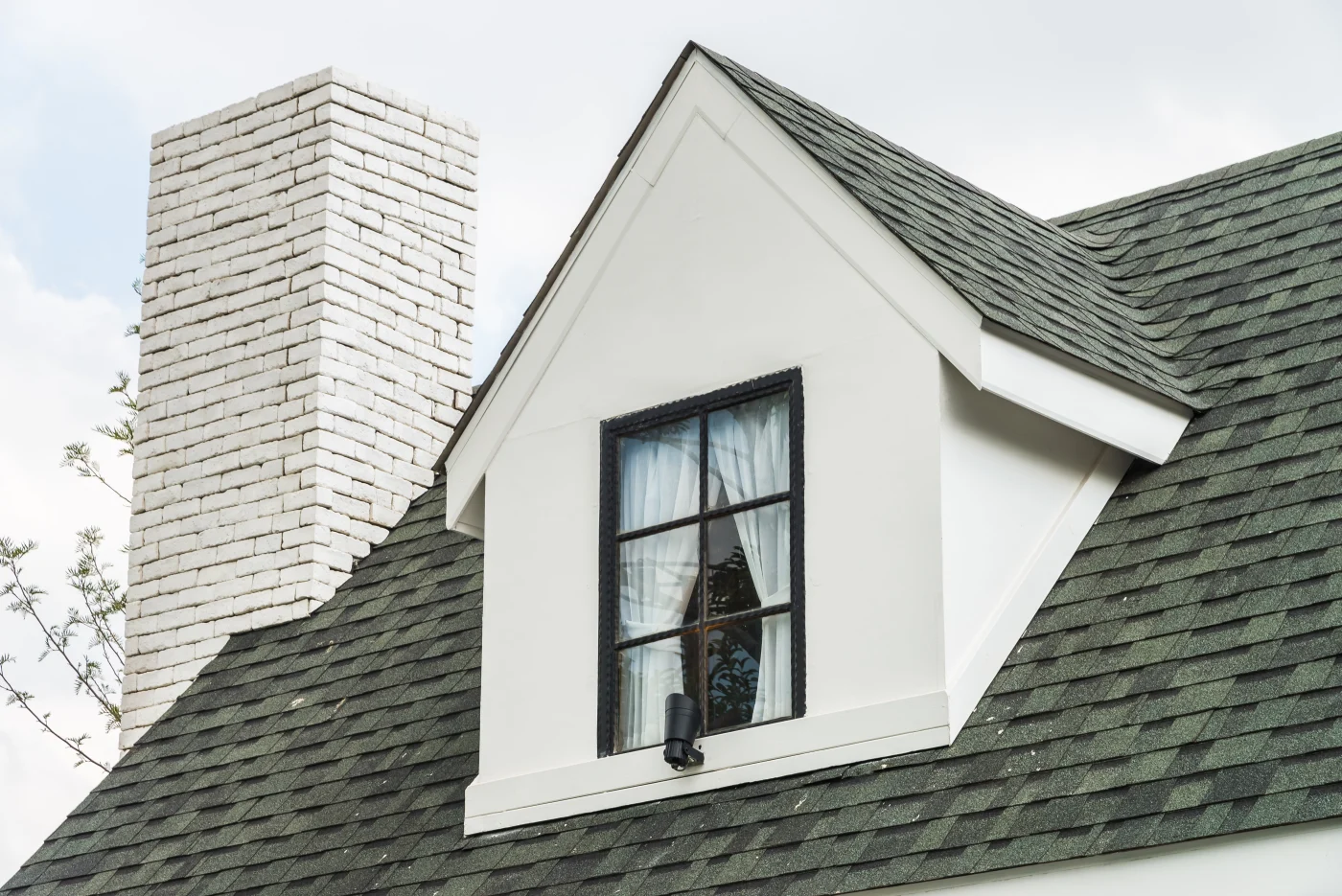 White dormer and painted brick chimney rising above a dark asphalt shingle roof on a restored home