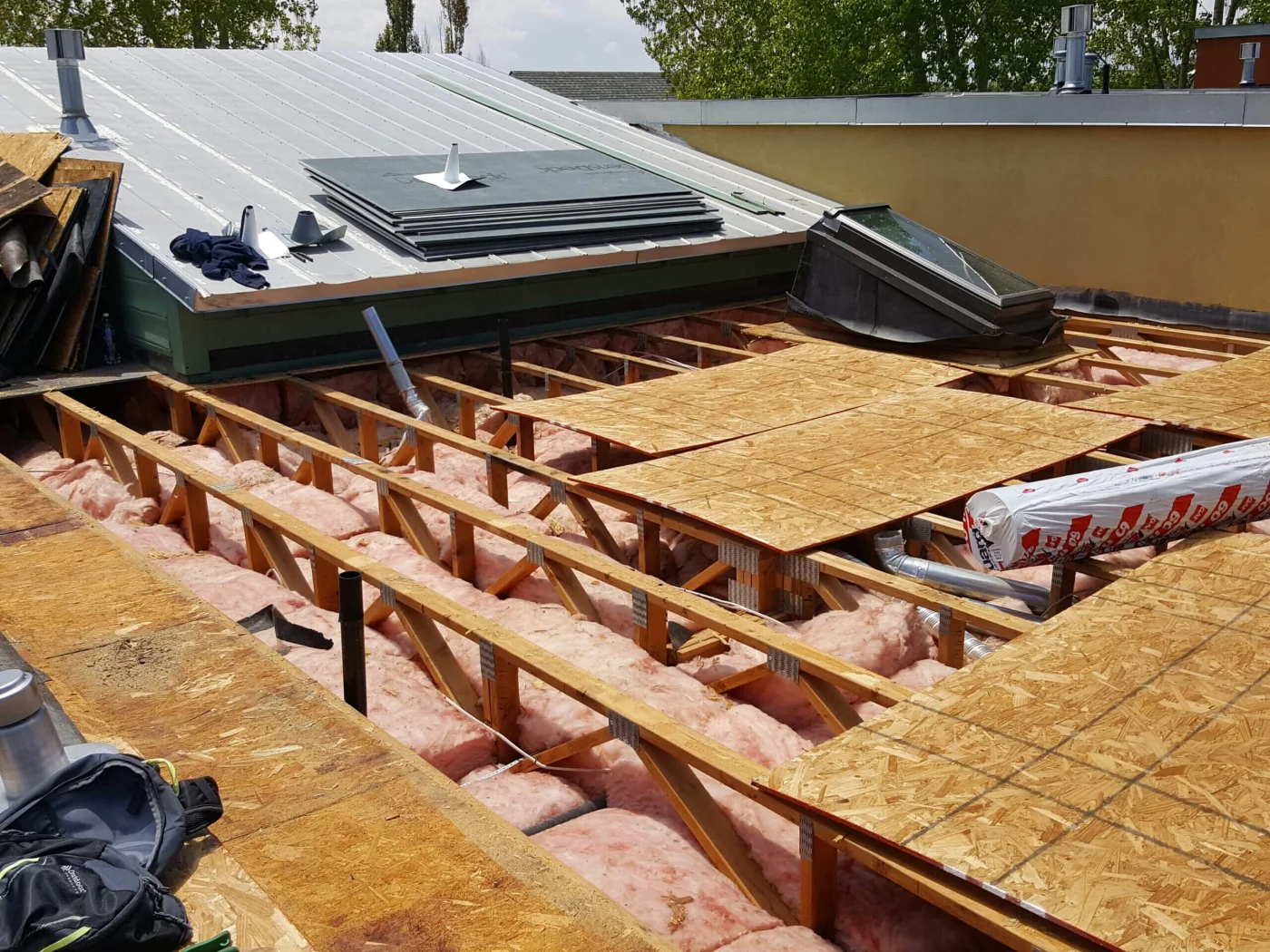 A roof reconstruction in progress for storm damage repair with exposed trusses, pink insulation, OSB decking, and nearby metal roofing materials
