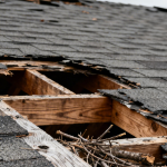 A storm-damaged asphalt shingle roof with a large hole exposing the wooden roof deck and broken debris inside.