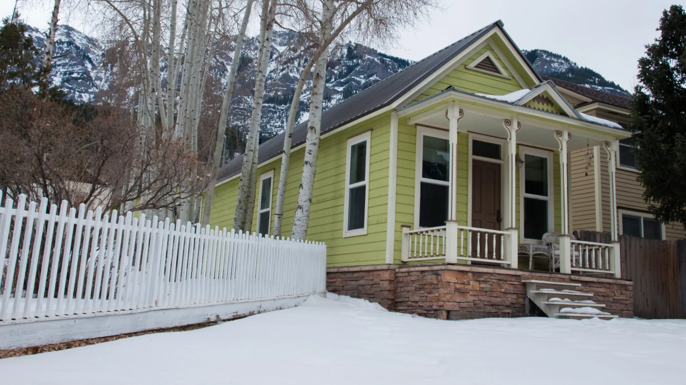 Small green cottage with front porch, white trim and fence, snow-covered yard, bare trees, and mountains in the background under a cloudy winter sky.