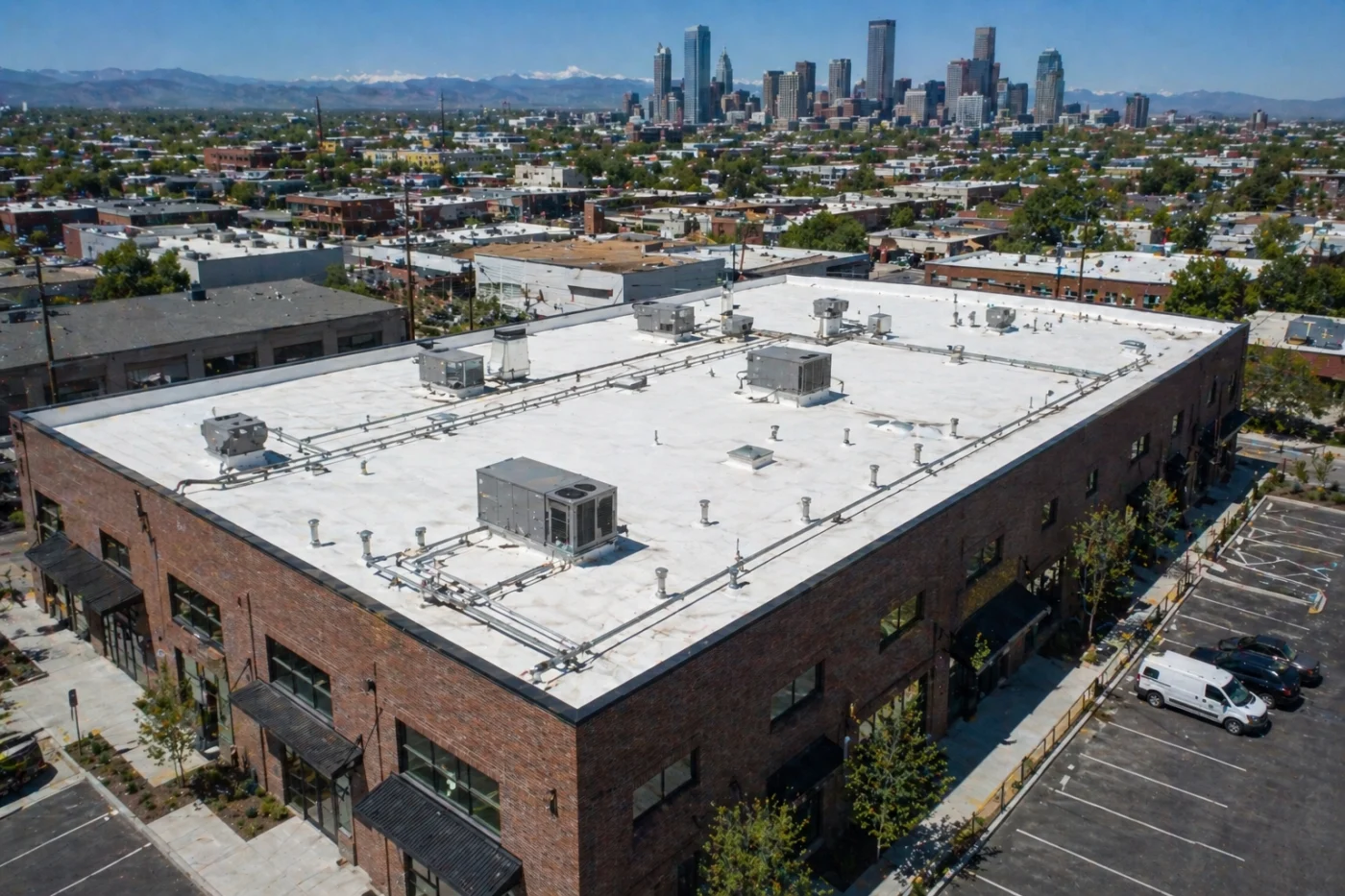 Angled aerial view of a Denver, CO commercial building with a white flat roof, rooftop HVAC units, brick exterior, skyline, and mountains.