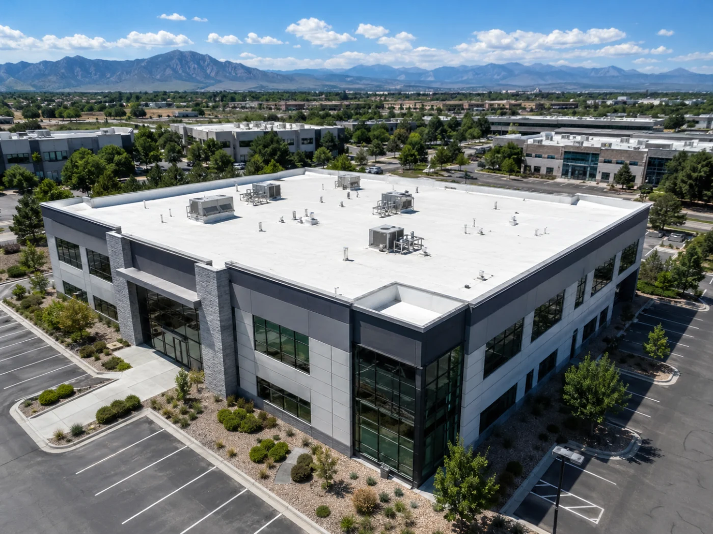 Angled aerial view of an Aurora, CO commercial building with a white flat roof, rooftop HVAC units, parking lot, and mountain backdrop.