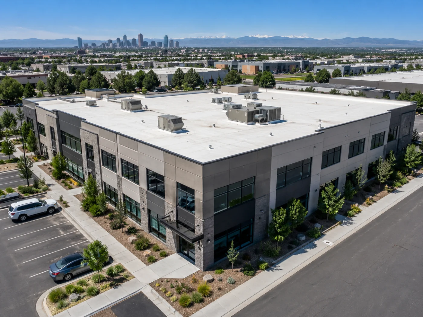 Angled aerial view of a Denver, CO commercial building with a white low-slope roof, rooftop HVAC units, parking lot, and city skyline.