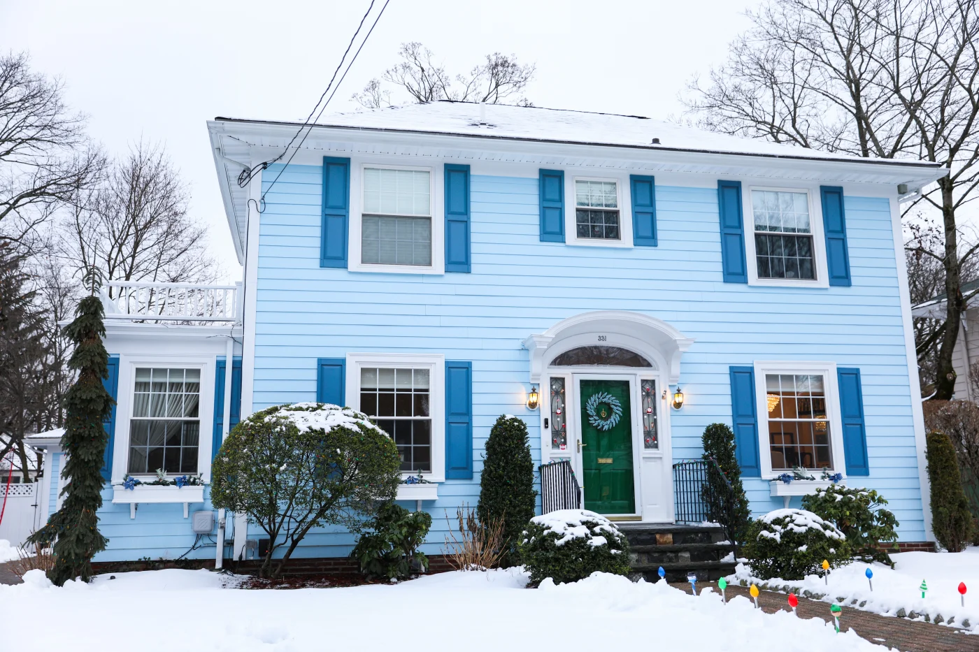 Two-story blue house with white trim and shutters, green front door, snow-covered yard, shrubs, and holiday lights in a winter residential setting.