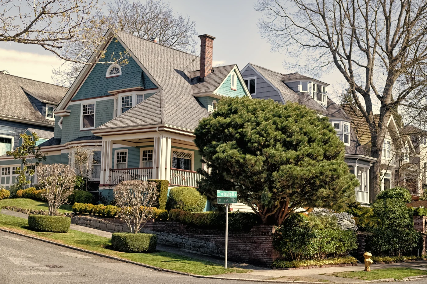 Colorful Craftsman-style home with blue-green siding, wraparound porch, brick chimney, manicured shrubs, and mature trees on a quiet residential street.