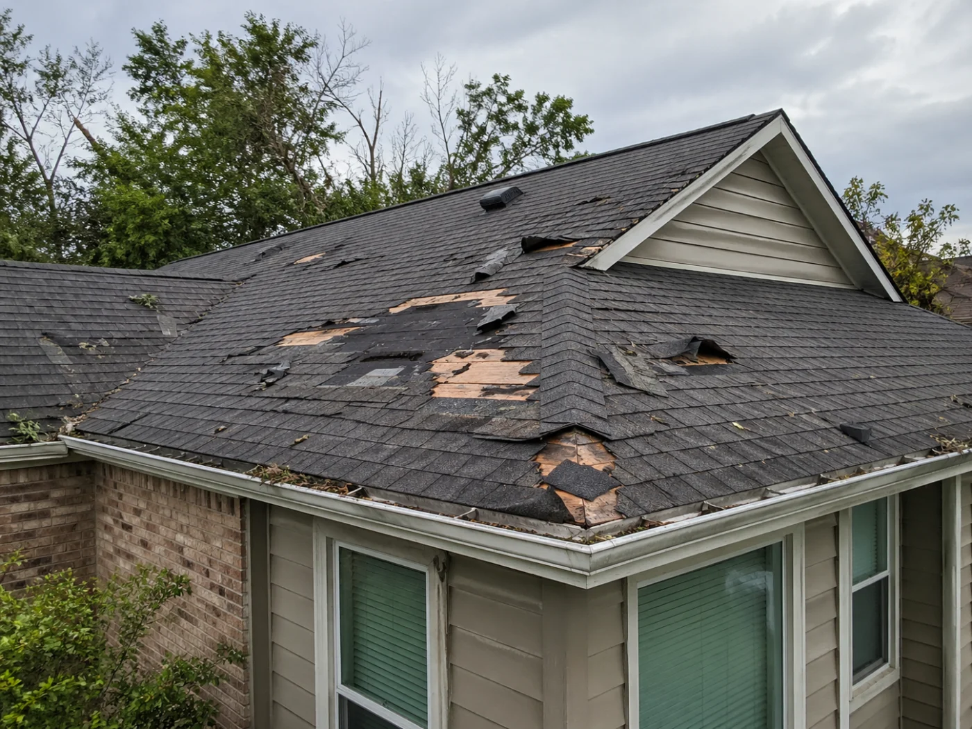 Storm-damaged home roof with torn shingles, exposed decking, debris-filled gutters, broken trees, and cloudy skies after severe weather.
