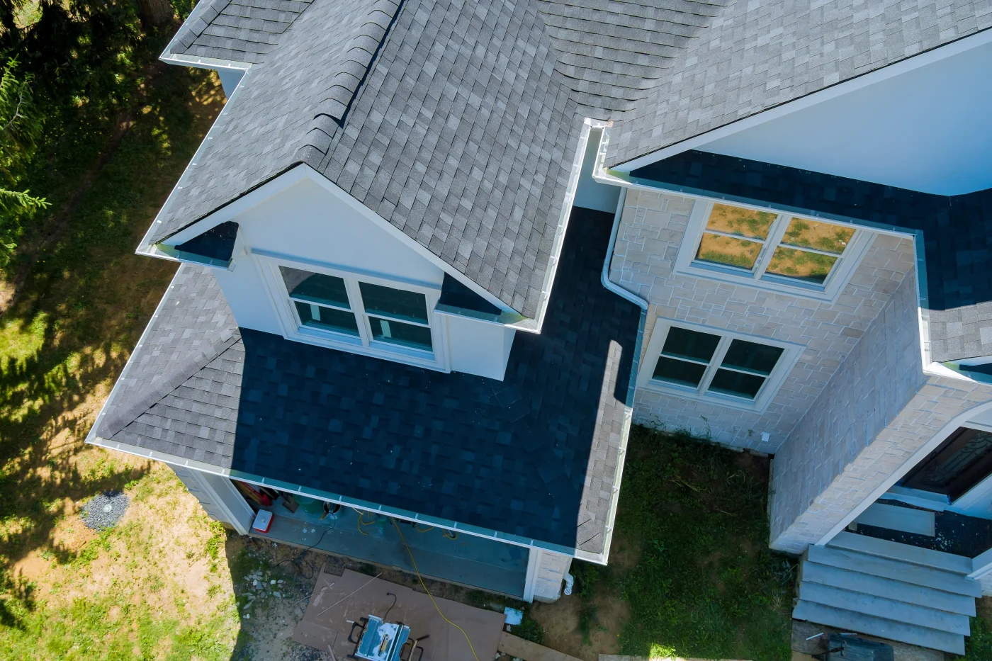 Aerial view of a gray shingle roof on a stone-and-white home, showing clean roof valleys, windows, front steps, and surrounding trees.