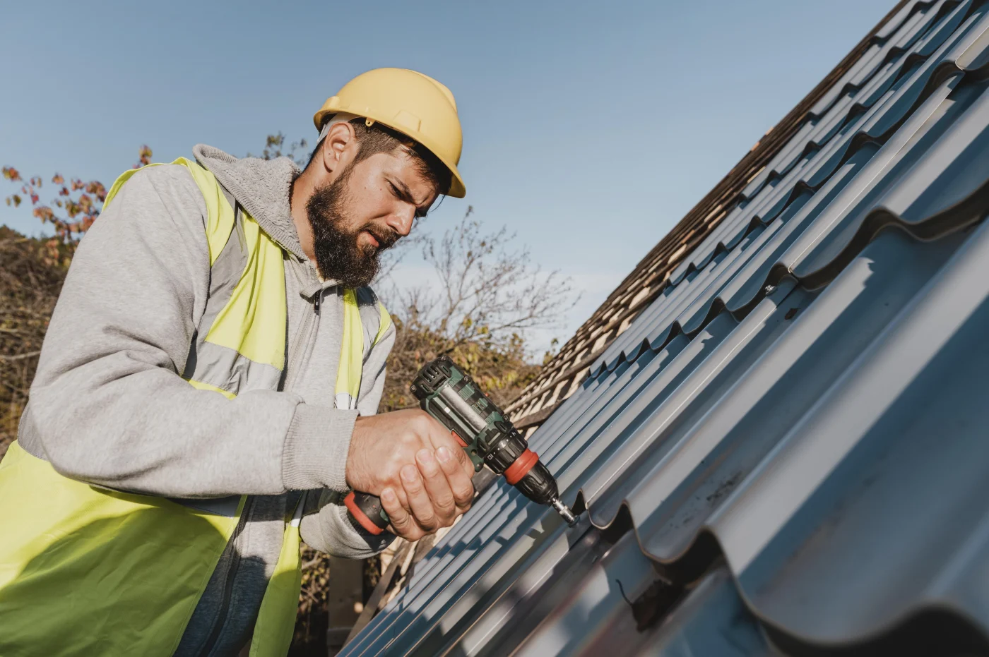Roofer in hard hat and safety vest uses a drill to fasten metal roofing panels on a sloped roof under a clear sky.
