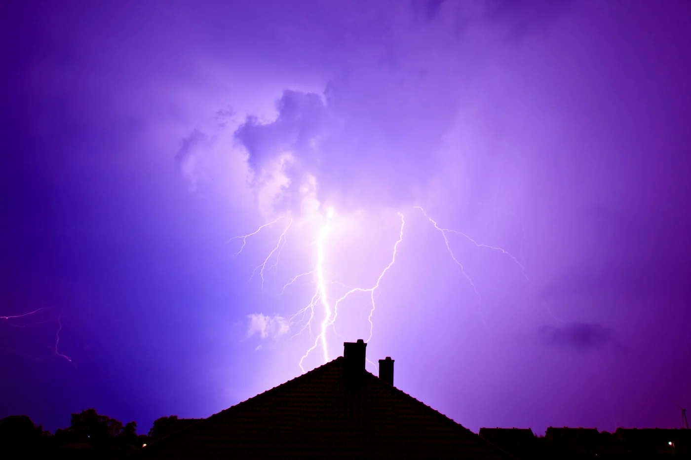 Lightning splits a purple storm sky above a dark roof silhouette, with bright flashes illuminating clouds over nearby homes.