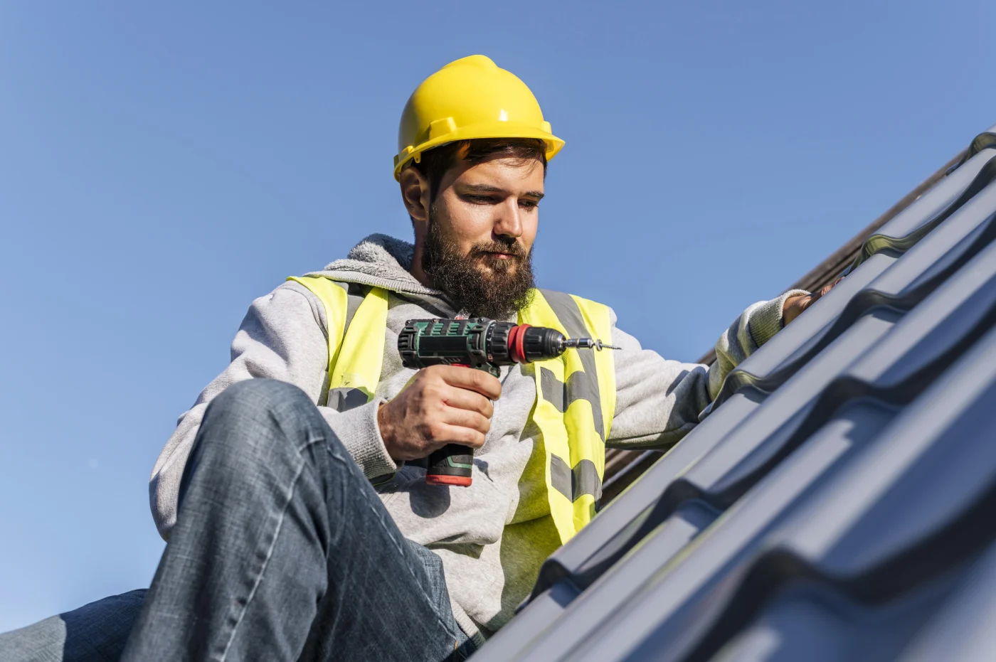 Roofer in a hard hat and safety vest holds a drill while working on metal roof panels under a clear blue sky.