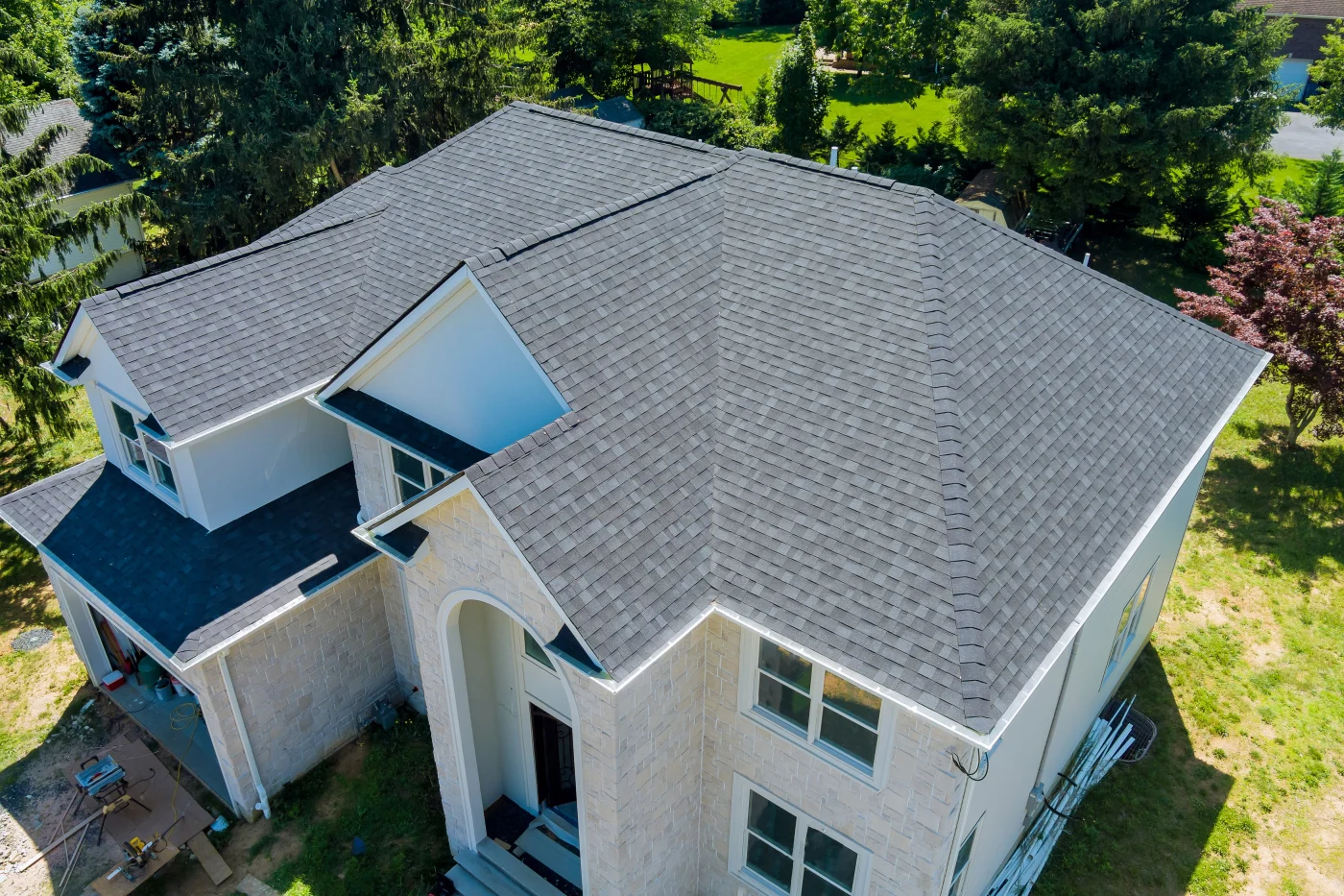 Gray shingle roof on a large stone-and-white home shown from above, with clean ridge lines, green trees, and roofing materials nearby.