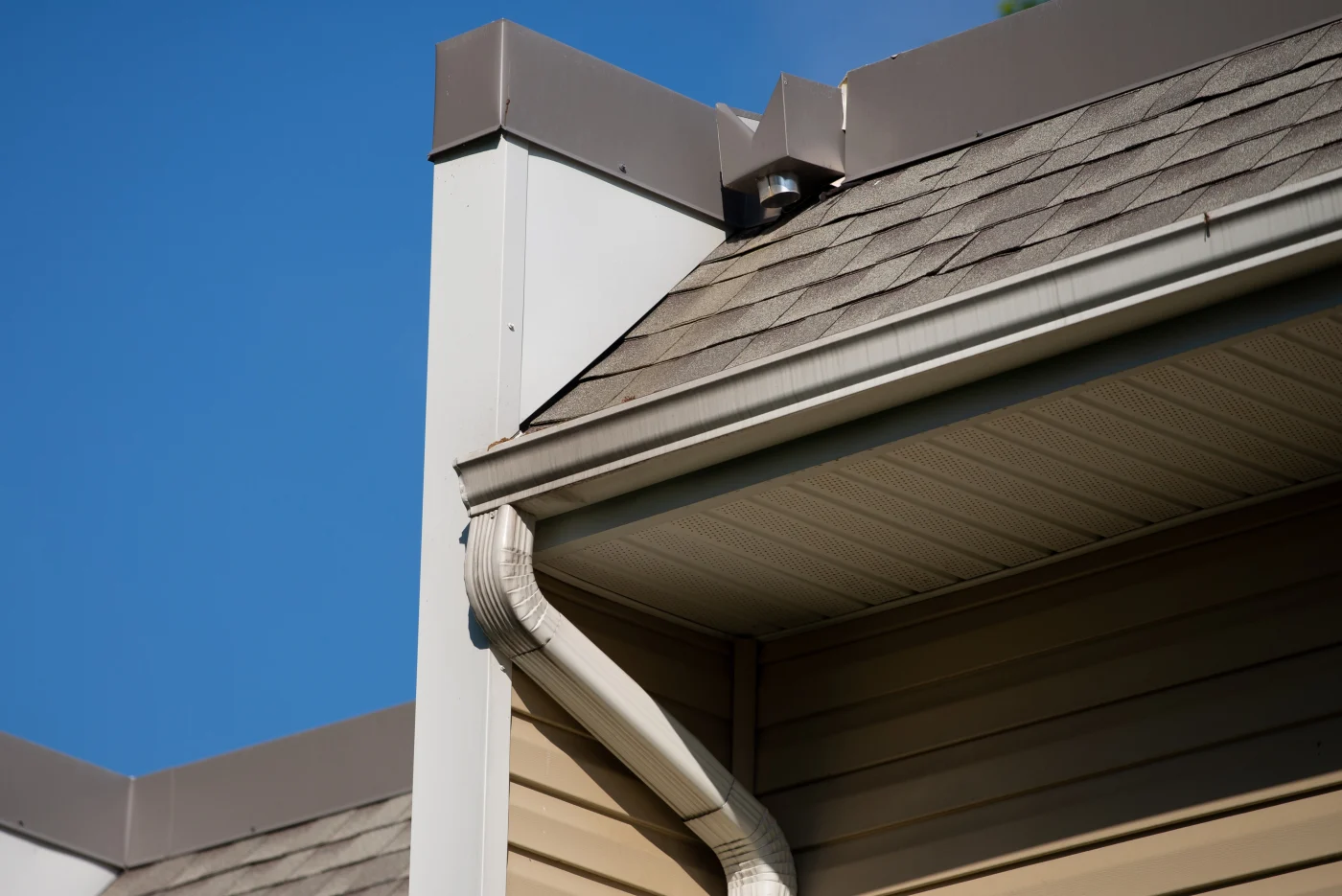 Residential gutter and downspout running along a shingled roofline with vented soffit panels and clear blue sky above.