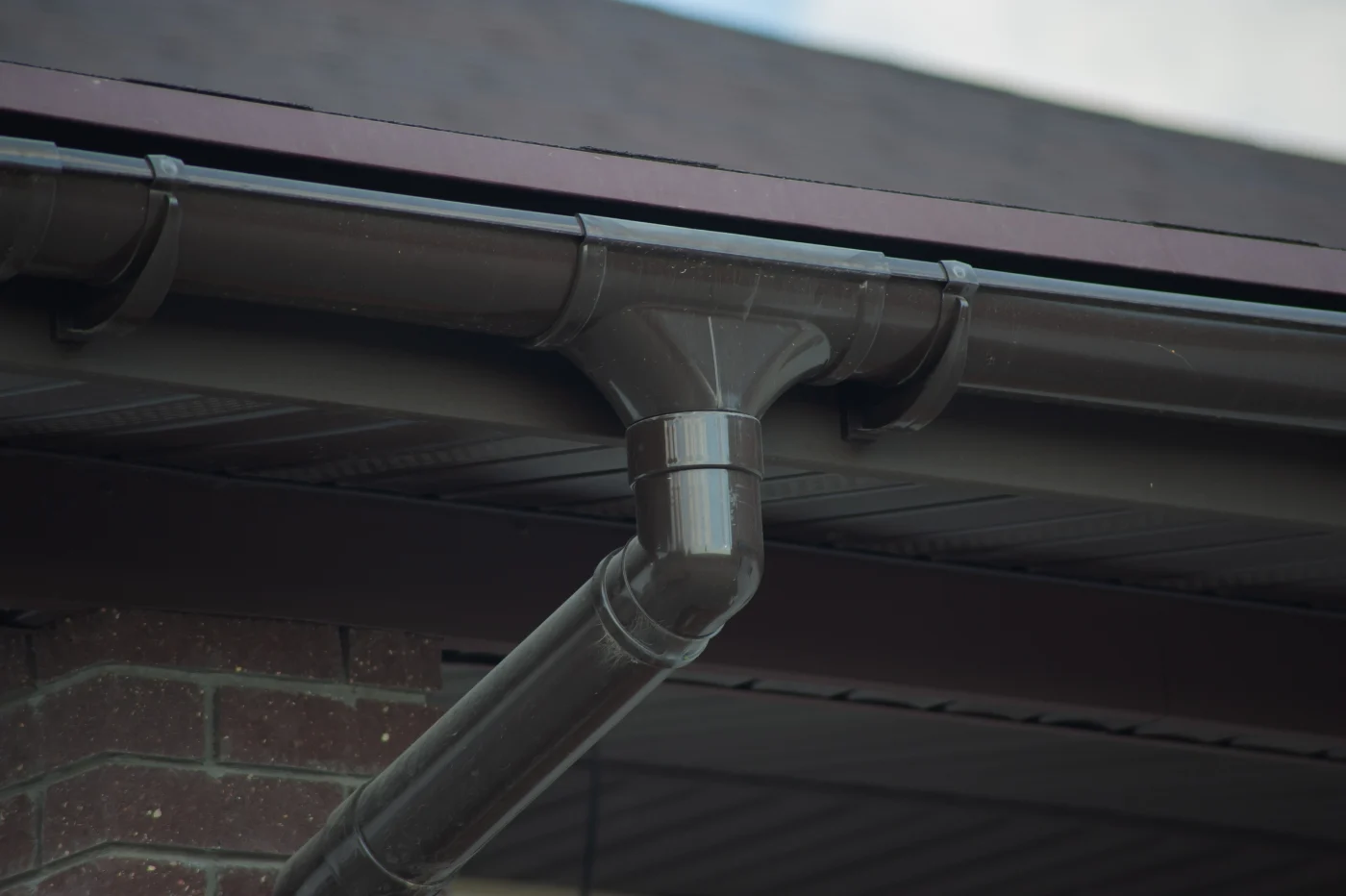 Black gutter and angled downspout attached to a roof edge, with dark soffit panels and brick siding beneath the eaves.