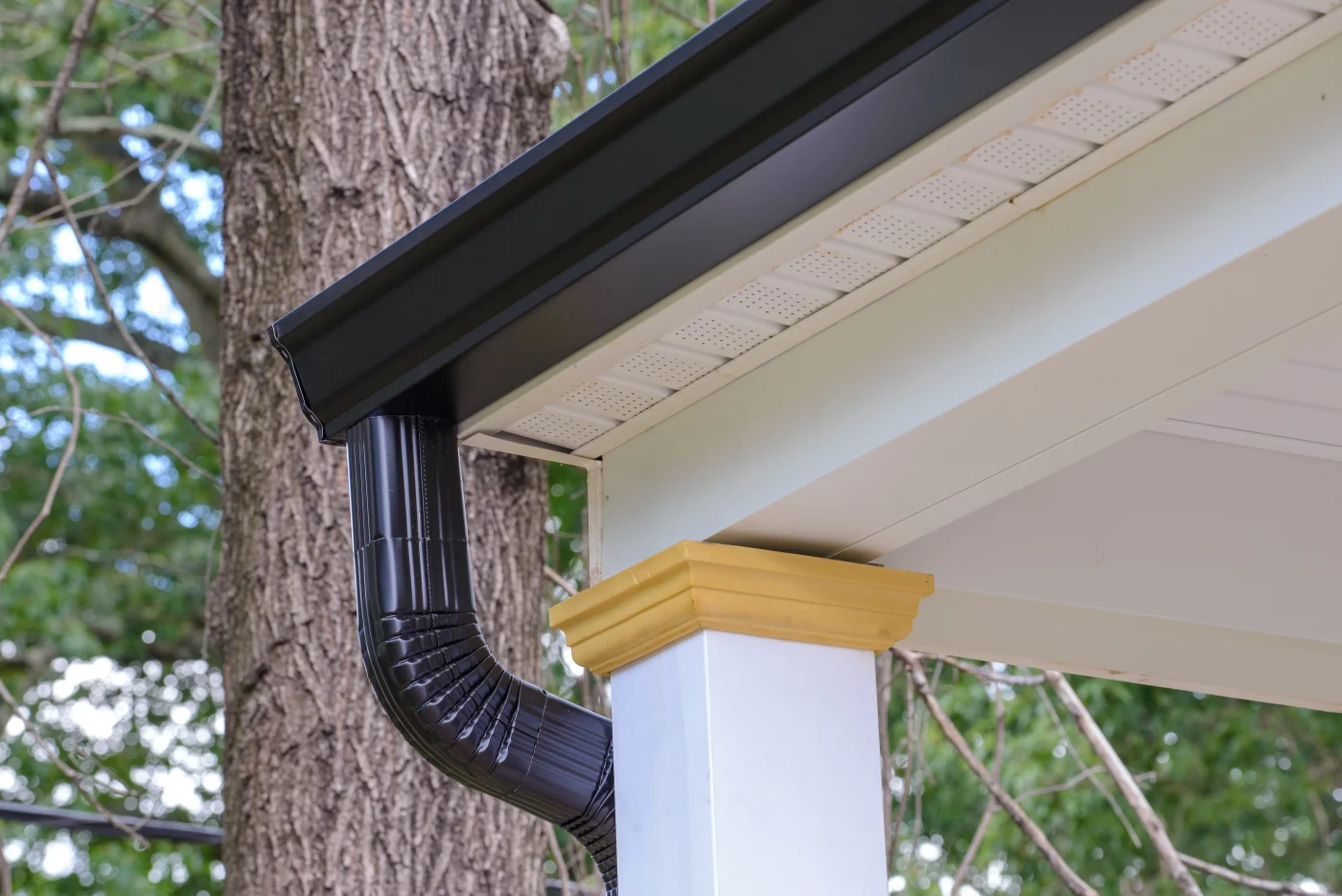Black residential gutter and downspout installed along a porch roof with vented soffit panels, white trim, and trees in the background.