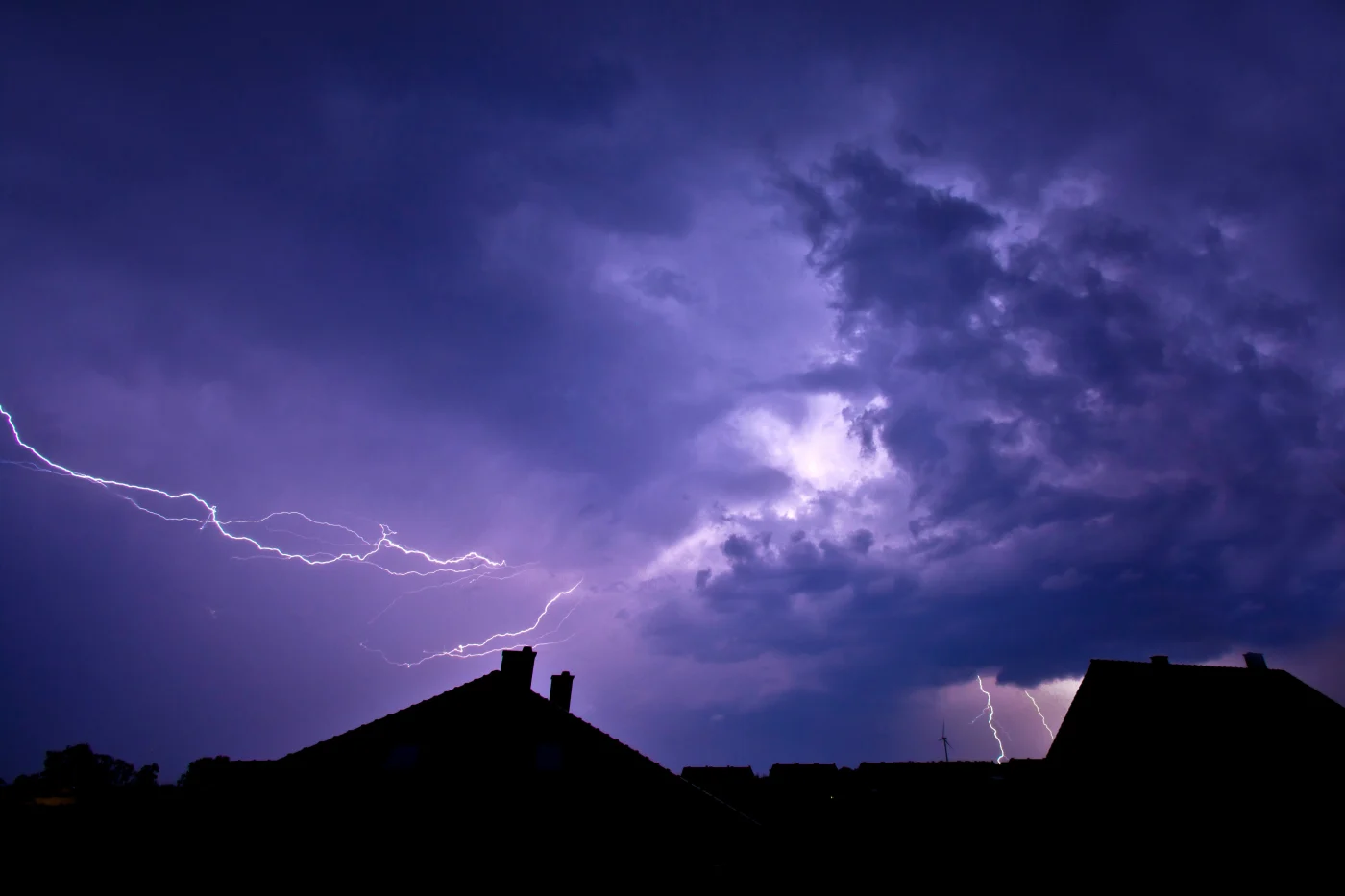 Lightning branches through dark purple storm clouds over silhouetted rooftops, with distant bolts illuminating the horizon.
