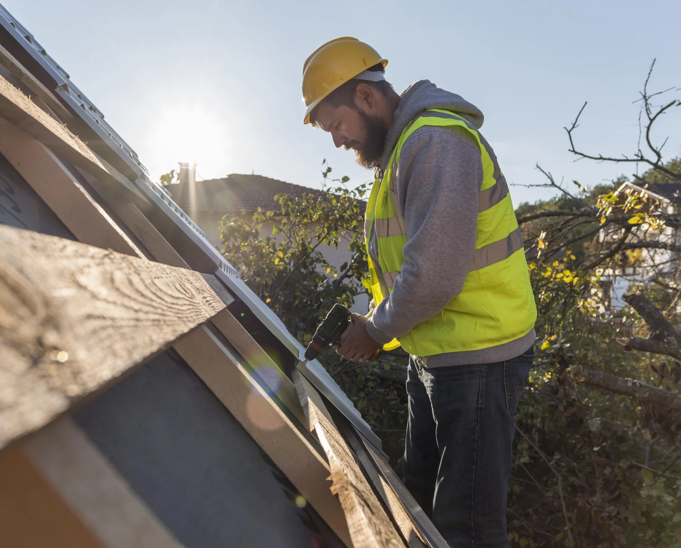 Roofer in hard hat and safety vest drills metal roof trim on a sloped roof, with sunlight, trees, and nearby homes behind him.