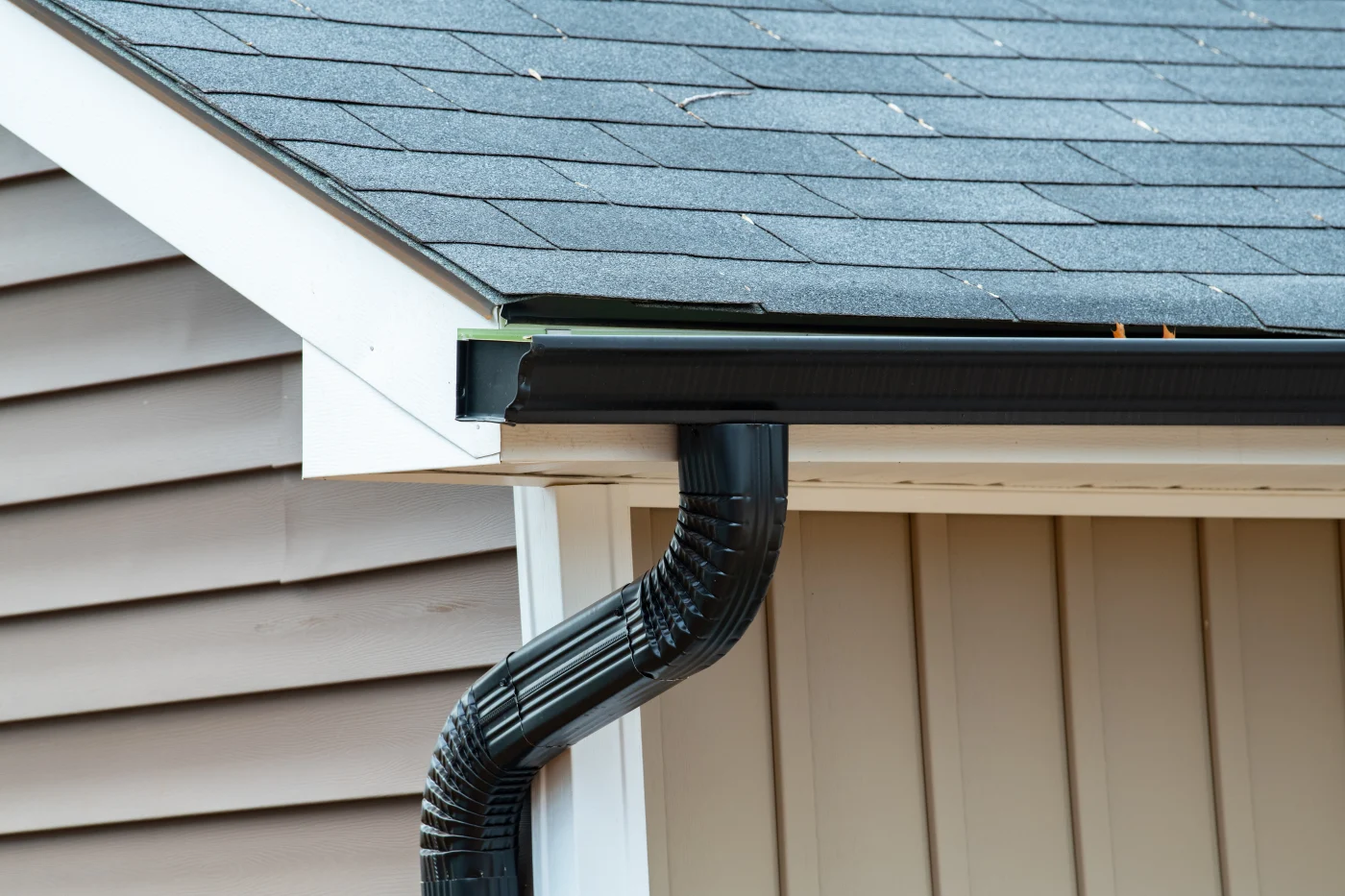 Black gutter and downspout installed along a gray shingle roof, with beige siding, white trim, and clean roof edge details.