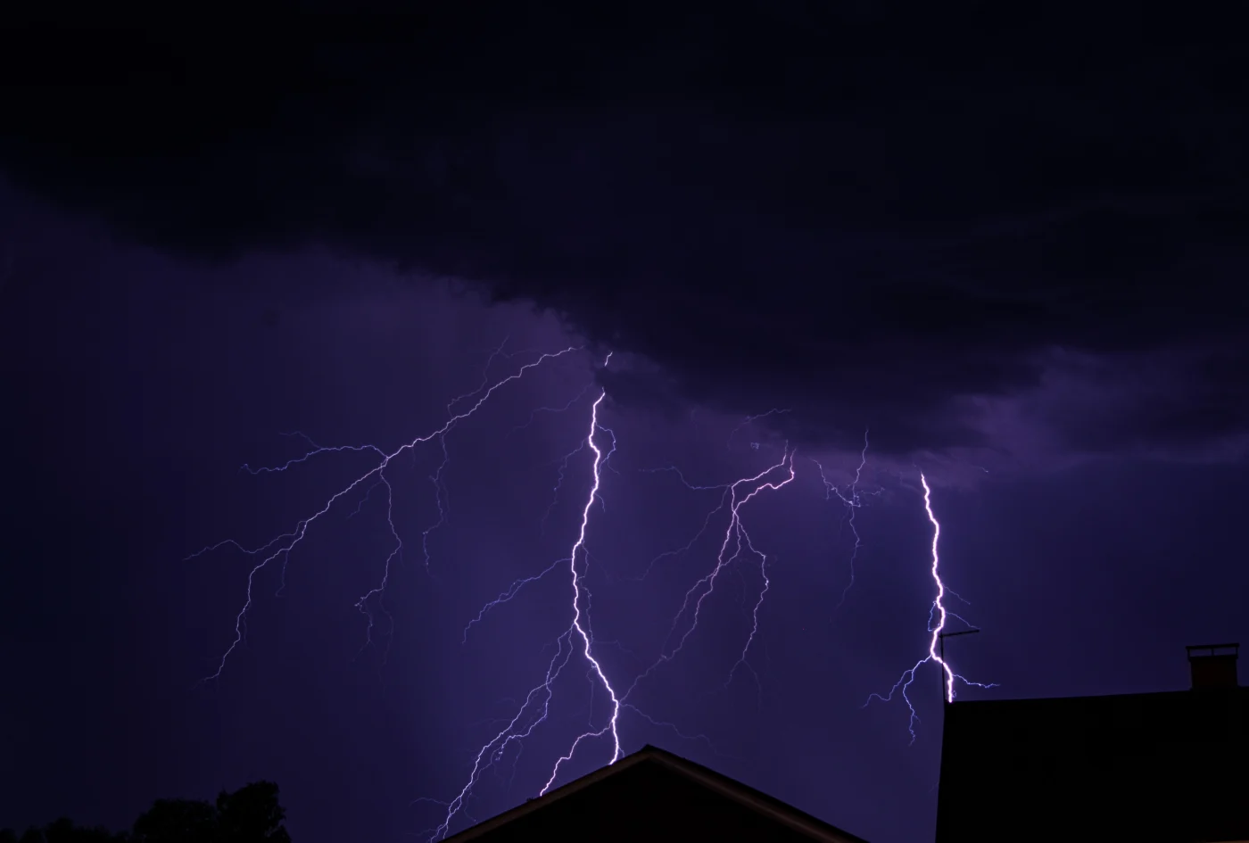 Lightning strikes through dark purple storm clouds above silhouetted rooftops, with bright bolts branching across the night sky.