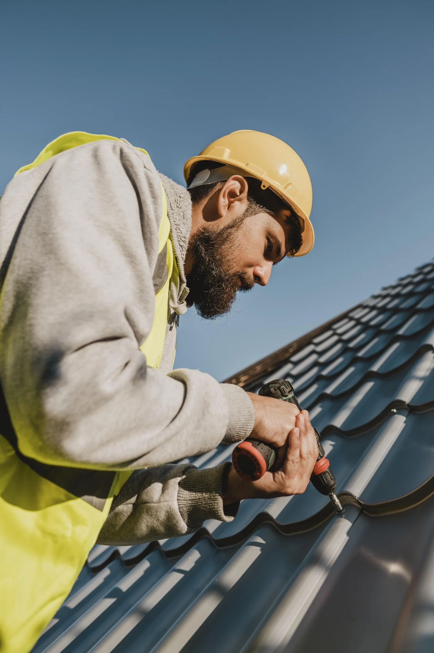 Roofer in hard hat and safety vest drills fasteners into metal roof panels on a sloped roof under a clear blue sky.