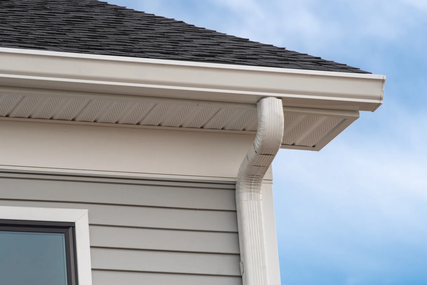 White residential gutter and downspout installed beneath dark roof shingles, with vented soffit panels and beige siding against a clear blue sky view.